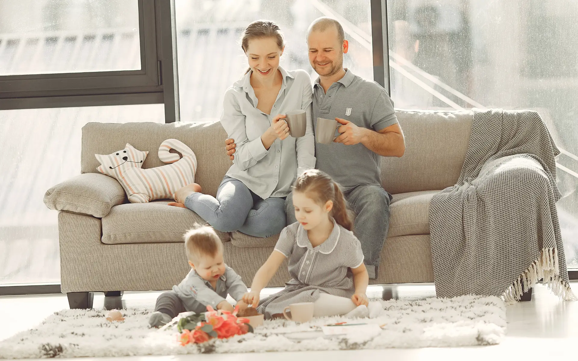 A happy family with two adults sitting on a sofa and two young children playing on the floor.
