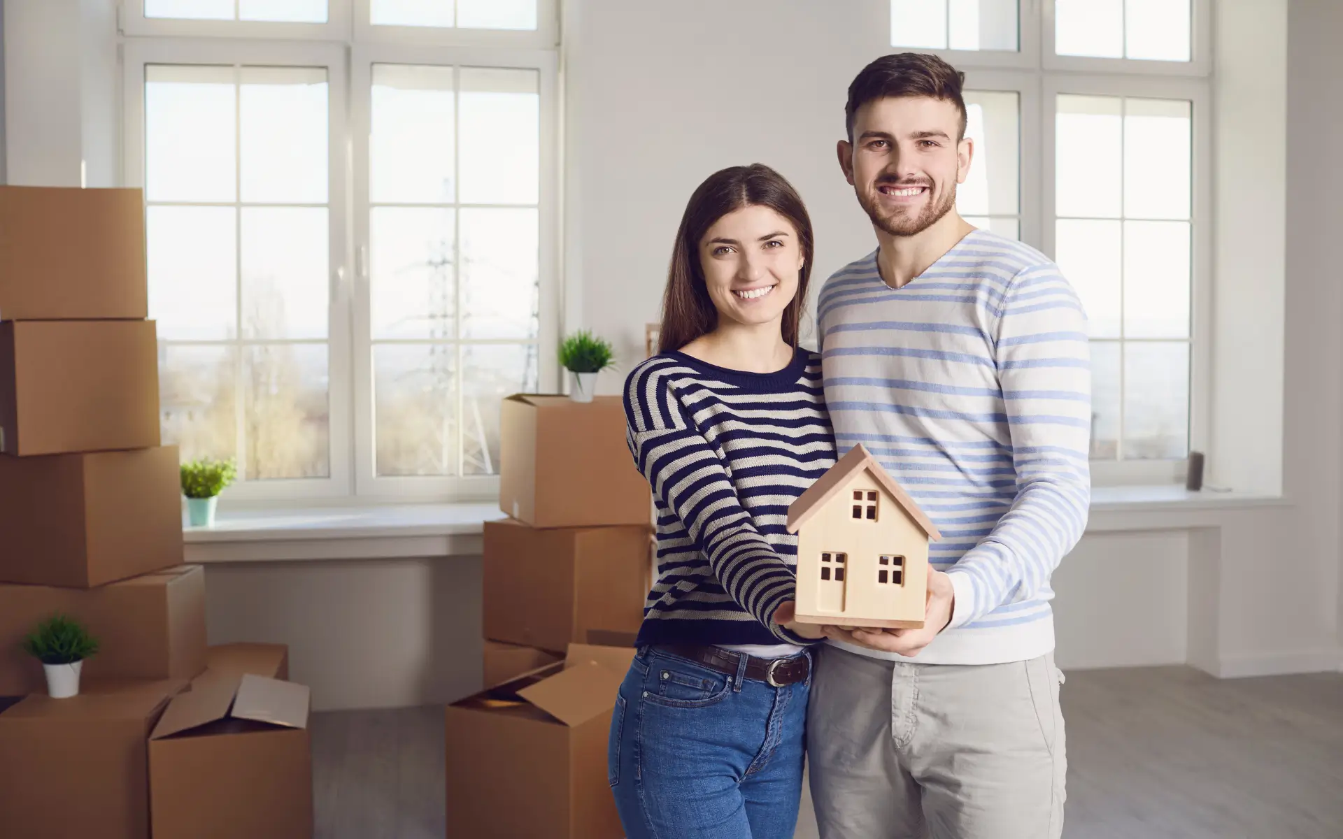 A smiling young couple stands amidst moving boxes, with the woman holding a small wooden house model.